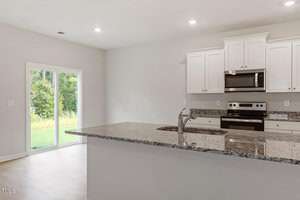 A modern kitchen with white cabinets and stainless steel appliances, including an oven and microwave. The granite countertop features a built-in sink. A sliding glass door opens to a lush green outdoor area, allowing natural light to illuminate the space.