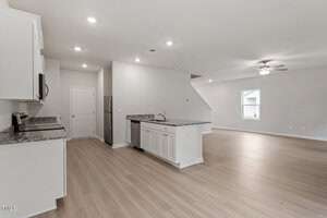 Spacious, modern open-concept kitchen and living room area with light wooden floors. The kitchen features granite countertops, white cabinets, and stainless steel appliances. Ceiling fan and window in the living area. Neutral color scheme throughout.