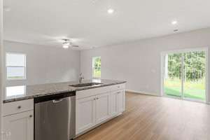 Modern kitchen with an island featuring a granite countertop and a built-in dishwasher. The open-concept space has light wood flooring, a ceiling fan, and large sliding glass doors offering a view of a grassy backyard and trees.