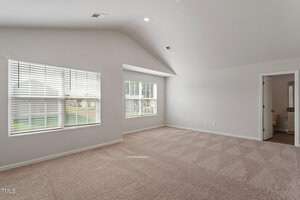 A spacious, carpeted room with high ceilings and neutral walls. Large windows on one side allow natural light to flood in. A small ensuite bathroom is visible in the corner, featuring a wooden door.