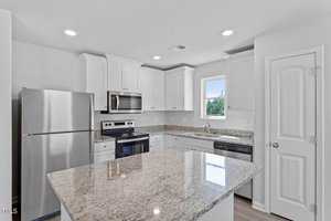 Modern kitchen with white cabinets, stainless steel appliances, and a granite island countertop. There's a window above the sink on the right, and recessed lighting brightens the space. A door is visible on the right side.