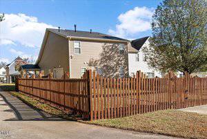 A two-story suburban house with beige siding and a black roof is surrounded by a brown wooden picket fence. The yard is grassy, and a large tree is visible. The sky is blue with scattered clouds.
