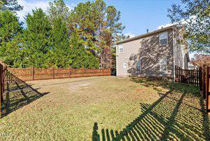 A fenced-in backyard with a grassy lawn, surrounded by tall trees. A two-story house with beige siding is on the right. The sky is clear and sunny, casting shadows on the ground.