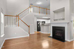 Spacious home interior with a wooden staircase, light-colored walls, and hardwood floors. The living room features a fireplace and large windows. The kitchen, equipped with modern appliances, is visible in the background.
