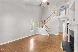 Spacious living room with high ceilings, light gray walls, and polished hardwood floors. A wooden staircase with white railings leads upstairs. Natural light enters through a small window. A white fireplace is on the right wall.