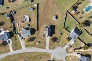 Aerial view of a suburban neighborhood showing several houses with driveways, yards, and trees. The image includes a curved road and a few fenced areas, with scattered small structures and a swimming pool.