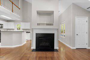 A spacious living room with high ceilings, featuring a central black fireplace with a white mantel. The room has wooden floors, light gray walls, and a kitchen area with white cabinets partially visible to the left.