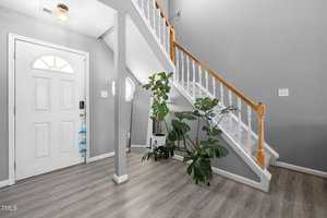 A modern home entryway featuring a light gray color scheme. A small staircase with white railings and wooden accents leads to the upper floor. Potted plants are placed at the base of the stairs, and a white door with a semi-circular window is on the left.