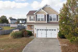 Two-story house with beige siding and brick accents, featuring a double garage door. A concrete driveway leads to the entrance. The yard is landscaped with grass and bushes, and a tree is on the right. Other houses are visible in the background under a cloudy sky.