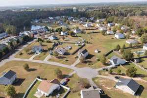 Aerial view of a suburban neighborhood with detached houses, large lawns, and a curving road. Trees are scattered throughout the area, and a water tower is visible in the distance.
