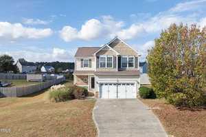 Nestled in the heart of Creedmoor, 2063 Covey Court is a charming two-story suburban house with a double garage, featuring beige siding and brick accents. The lawn is partially dry, complemented by a tree on the right and a fenced backyard. The sky is partly cloudy, with neighboring houses visible beyond.