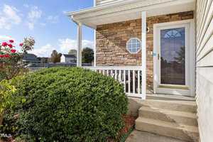 A house entrance with a stone accent wall and white railing. A glass door leads inside, with trimmed shrubs and red flowers adorning the pathway. A blue sky with clouds is visible in the background.
