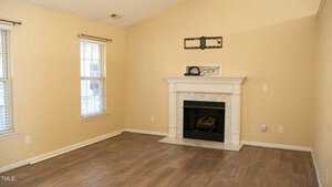 A vacant room with cream-colored walls and wooden flooring. It has a marble fireplace centered on one wall and two tall windows on the left. A TV mount is installed above the fireplace.