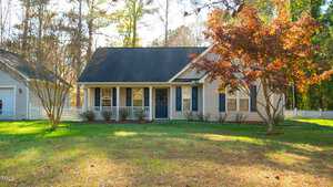 A quaint suburban house with a dark roof and light siding is surrounded by trees. The front yard has a few bare trees, and a vibrant red-leaved tree. A white picket fence runs along the property, and the grass is lush and green.