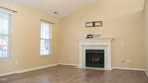 A vacant living room features light beige walls, two tall windows with blinds, and a white fireplace with a gray stone surround. The floor is wood laminate. A wall mount for a TV is placed above the fireplace. Natural light filters in through the windows.