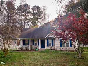 Located at 4040 Old Franklinton Road, this charming single-story house in Franklinton features beige siding, blue shutters, and a dark roof. A front porch overlooks the well-kept lawn and red-leaved tree, while a white picket fence peeks through the tall trees in the background.