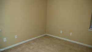 An empty room with beige walls and carpet. The room features several electrical outlets on the walls, and a partially visible window with blinds on the right. The baseboard is white, contrasting the neutral tones of the room.