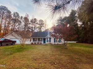 A single-story house with a porch sits amidst a grassy yard. A red truck is parked on the left side. Tall trees with autumn leaves surround the property. The sky is partly cloudy.