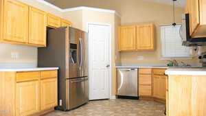 A kitchen with light wooden cabinets, stainless steel appliances including a refrigerator and dishwasher, a small window with blinds, and beige walls. The flooring is a patterned tile, and there is a door leading to another room.