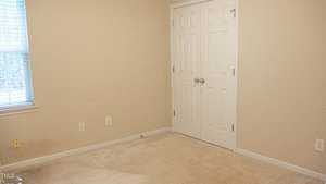 A small, empty room with beige walls and carpet. There is a closed white double door closet on the right and a window with blinds on the left. The room is well-lit and has electrical outlets visible on the walls.