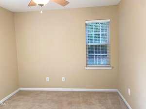 An empty room with beige walls and carpet, featuring a window with white blinds. A ceiling fan is installed above, and there are electrical outlets on one wall.