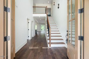 Spacious interior of a modern home with wooden floors and a staircase leading to an upper level. Glass doors open into the bright hallway, and natural light floods the space from windows and a balcony railing above.
