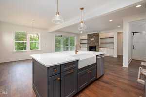 Modern kitchen with a large white countertop island, dark cabinetry, and brass fixtures. Open floor plan leads to a living area with a stone fireplace and built-in shelves. Sliding glass doors overlook a green backyard. Hardwood floors throughout.