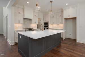 A modern kitchen featuring a large island with a white countertop and dark base. The background includes white cabinets, a stainless steel oven, a gas stove, and two pendant lights hanging over the island. Dark hardwood floors complete the look.