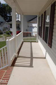 A covered porch with a white railing and brick trim. The porch leads to the front yard, with neighboring houses visible in the background. Sunlight casts shadows of the railing onto the concrete floor.