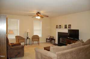 A living room with beige walls and carpet, featuring a ceiling fan, two windows with blinds, a sofa, an armchair, a recliner, and a wooden coffee table. A fireplace with family photos on the mantel and a flat-screen TV are present.