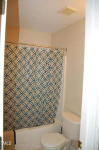 Bathroom with a shower-tub combo, featuring a blue and white geometric patterned curtain. A white toilet with the seat down is in the foreground. The walls are light-colored, and a small portion of a towel is visible hanging near the tub.