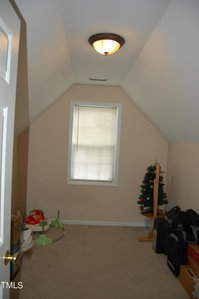 A small attic room with sloped ceilings, a single window with blinds, and a ceiling light. The room contains a small decorated Christmas tree, a few boxes, and some scattered children's toys.