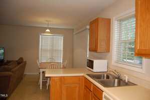 A kitchen with wooden cabinets, a double sink, and a white microwave on the counter. A dining area with a wooden table and chairs is near a window with blinds. A brown couch is visible in the living room area.