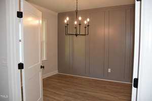 A small, empty room with light-colored hardwood flooring and a gray accent wall featuring vertical paneling. A modern chandelier with exposed bulbs hangs from the ceiling. A white door is partially open, and a window is visible on the left.