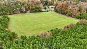 Aerial view of a large grassy field surrounded by dense trees with autumn foliage. In the background, there's a small road and a few residential buildings. The scene suggests a rural setting with a mix of green, orange, and brown hues.