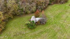 Aerial view of an abandoned house surrounded by overgrown vegetation in a grassy field. The roof is rusted and partially collapsed. There are dense trees in the background.