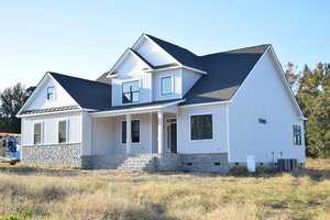A modern, two-story white house with gray stone accents, black roof, and covered front porch, set in a grassy field under a clear blue sky.