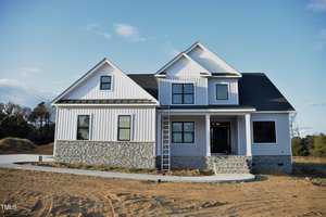 Nestled in Franklinton, the newly constructed two-story house at 4301 Bragg Farm Lane showcases white siding and a dark roof. Its stone facade graces the lower half, while a ladder leans against the front. The front yard is unlandscaped with bare dirt and a paved driveway framed by trees in the background.