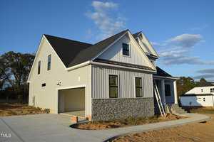 A newly constructed two-story house with light gray siding and a stone facade. The driveway is unfinished, and an extension ladder leans against the front. The sky is blue with scattered clouds, and trees are visible in the background.