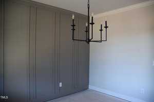 A room with light gray walls, featuring a modern black chandelier with six candles and exposed bulbs. The floor is carpeted, and a section of the wall has decorative paneling.