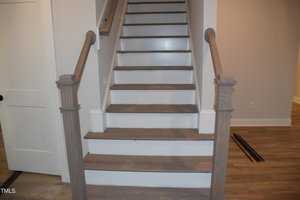 A wooden staircase with brown steps and white risers is shown. It has matching brown handrails on both sides. The staircase is set against a light-colored wall, and there is a closed white door on the left. Some wooden planks are on the floor.