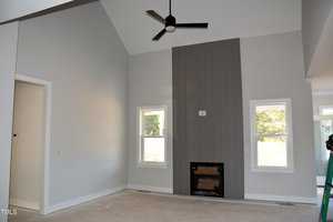 A room with high vaulted ceilings features a central fireplace beneath a gray accent wall. Two windows flank the fireplace, allowing natural light to enter. The flooring is unfinished, and a black ceiling fan hangs from above.