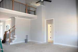 A room with high ceilings and gray walls, featuring an unfinished floor. There's a staircase with wooden railings leading to a loft area. A ceiling fan is visible, and several doors are along the walls. Natural light enters through nearby windows.