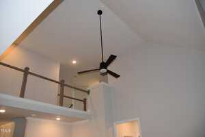 A modern ceiling fan with a circular light fixture is mounted on a high vaulted ceiling. The walls are painted light gray, and recessed lights are embedded in the ceiling. A wooden railing runs along an upper ledge.