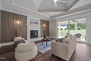 A modern living room features a ceiling fan and recessed lighting, with a white fireplace against a paneled wall. Two beige chairs and a sofa surround a small table on a patterned rug. Sliding glass doors open to a patio with outdoor seating and greenery.