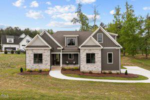 Located at 2504 Jessis Court in Franklinton, this modern suburban house boasts a striking mix of stone and dark siding exterior. It features a gabled roof and a covered front porch. A curved driveway, set amidst a spacious yard with trees, leads to the neatly landscaped lawn and young shrubs.