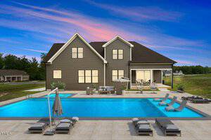 A modern two-story house with a spacious backyard features a large rectangular swimming pool. Several lounge chairs and umbrellas are set around the pool on a tiled patio, and a covered patio area is visible to the right. The sky shows a sunset.