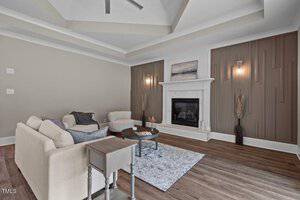 A modern living room with a beige sofa and armchair, a small table, and a light-colored rug on wooden flooring. The room features a fireplace with a painting above it, decorative wall paneling, and two wall sconces. Ceiling has recessed design.