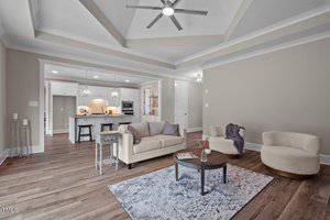 A modern living room with a beige sofa, two round chairs, and a small coffee table on a gray rug. The room features a tray ceiling with a ceiling fan. In the background, there's a kitchen with white cabinets and a bar area with stools.