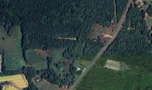 An aerial view of the rural Roxboro landscape reveals dense green forests, a pond, and open fields crisscrossed by NC Hwy 49. The road cuts diagonally, passing several structures scattered over both cleared and wooded land near 9515 NC 49.